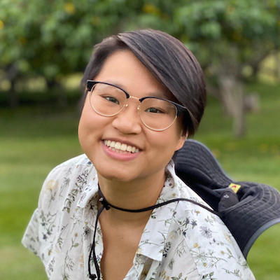 Connie Chang, a queer trans Chinese-American actual play GM and TTRPG designer smiles at the camera. They are wearing fashionable horn-rimmed-style glasses, a bucket hat tied around their shoulders, and a botanically-themed short sleeved button down.