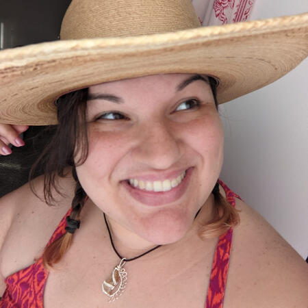 Mo, a Latine TTRPG content creator, writer, and sensitivity consultant. They are smiling and looking off to the right of the camera wearing a straw hat, pigtails, and an intricate silver pendant.